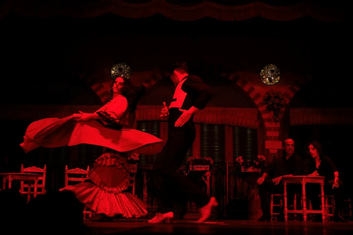 A woman in a red ruffled skirt and a man in black and red spin on a dark stage in front of an arched, patterned wall with small wooden tables and two seated onlookers