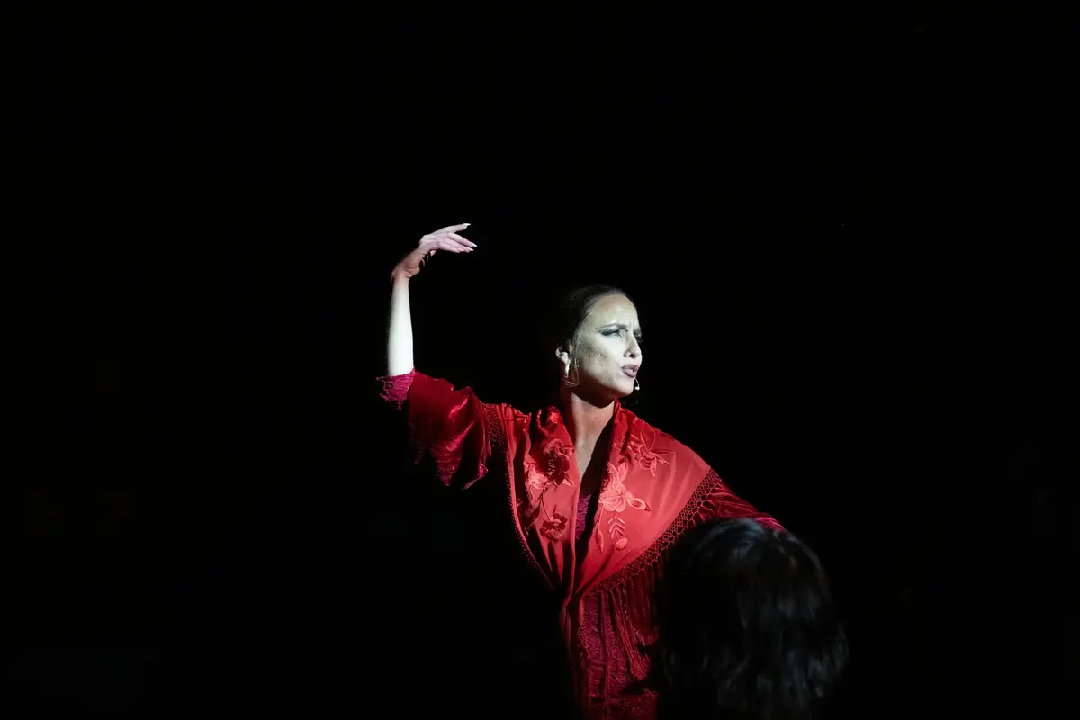 A dancer in a red embroidered shawl raises one arm in a single spotlight against a black background
