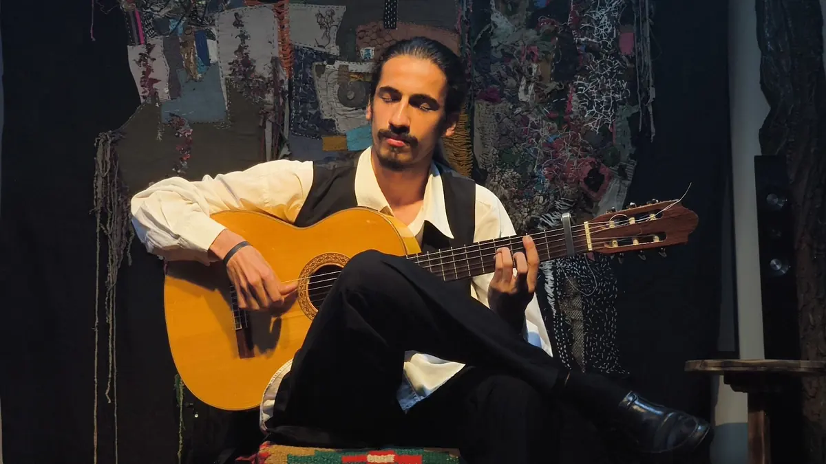 A flamenco guitarist in a white shirt and black waistcoat sits with one leg crossed over the other and the guitar held nearly flat, playing in front of a patterned cloth backdrop